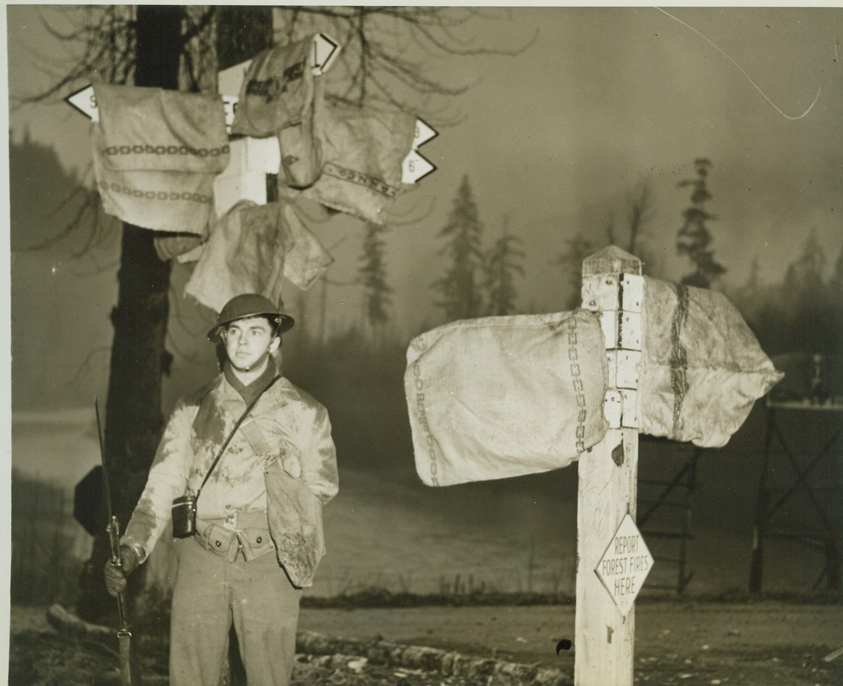 It’s _______ Miles to ________., 2/7/1942. One thing you can’t show in a photograph is the location of U.S. troops. That’s why these signposts are covered as a soldier stands sentry somewhere in the Pacific Northwest. As you can see from the signs, it’s several miles to some place or other. Anyway, it’s rainy there. Or was when the picture was taken. Credit: ACME.;
