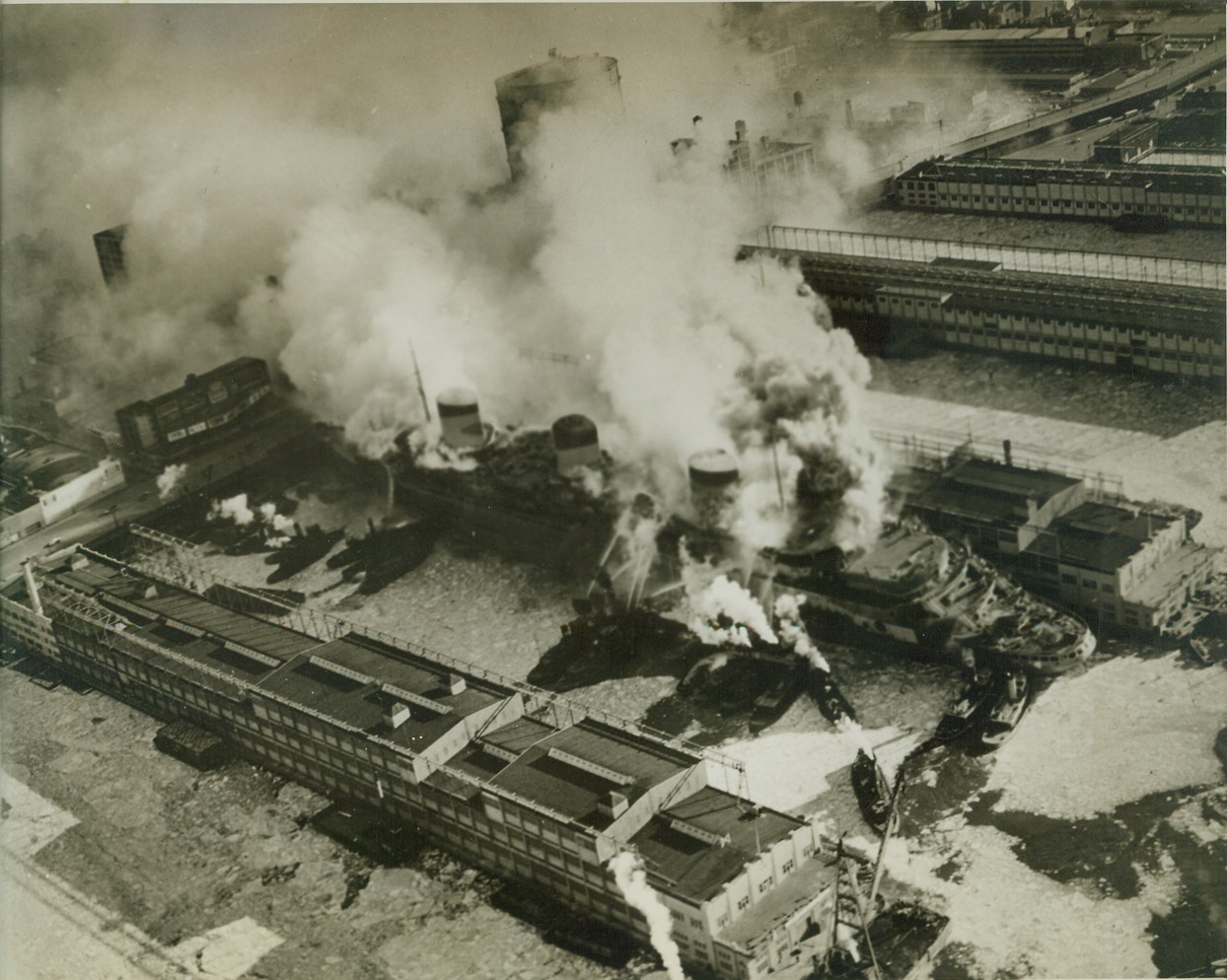 U.S.S. LAFAYETTE BURNS AT PIER, 2/9/1942. NEW YORK, N.Y.—Smoke billowing up both fore and aft from flames which gutted her upper decks, the former French luxury liner Normandie-now the U.S.S. Lafayette-burns with loss estimated unofficially at five million dollars at her Hudson River Pier. At lest one man, a workman trapped aboard the ship, was dead and at least 75 others injured. Photo made from a U.S. Army plane, shows fireboats playing streams of icy water on the ship, while city firemen on the dock attacked the flames on the starboard side. Credit: OWI Radiophoto from U.S. Army by 2nd Air Base Squadron, ACME;