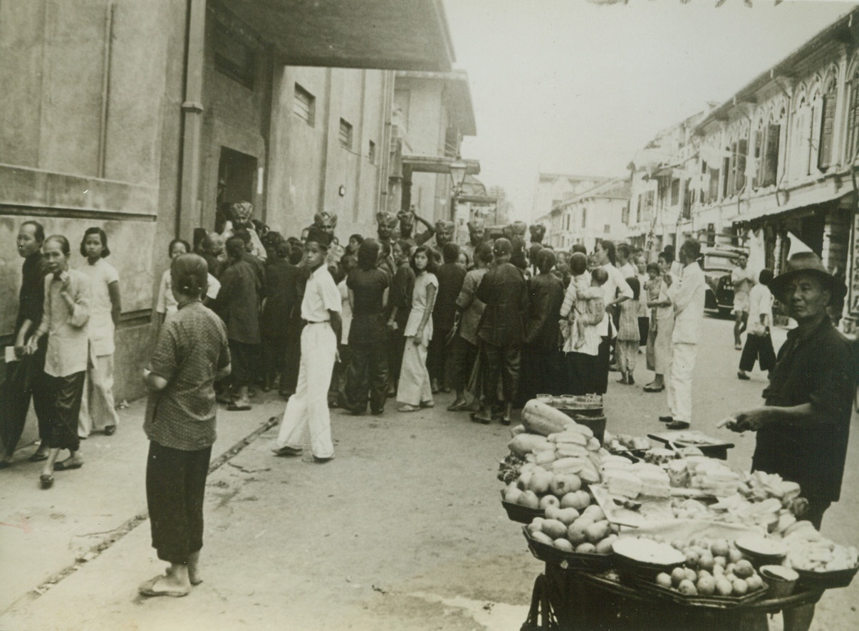 Security Registration in Singapore, 2/6/1942. Singapore – For a long time the people of Singapore have recognized the Jap threat to that great naval base.  Here, weeks before the start of the siege of Singapore, residents of the city’s Chinese quarter are shown crowding into a building for security registration.  A Chinese hawker peddles Malayan fruits in the street. Credit line (ACME);