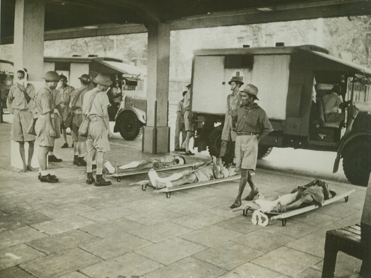 Malayan Chinese Aid British Wounded, 2/11/1942. Malaya – Chinese stretcher bearers have put wounded British soldiers down outside a Malayan station, while they wait for loaded ambulances to move on.  Other ambulances are coming up and the Chinese watch their patients while they wait. Credit line (ACME);