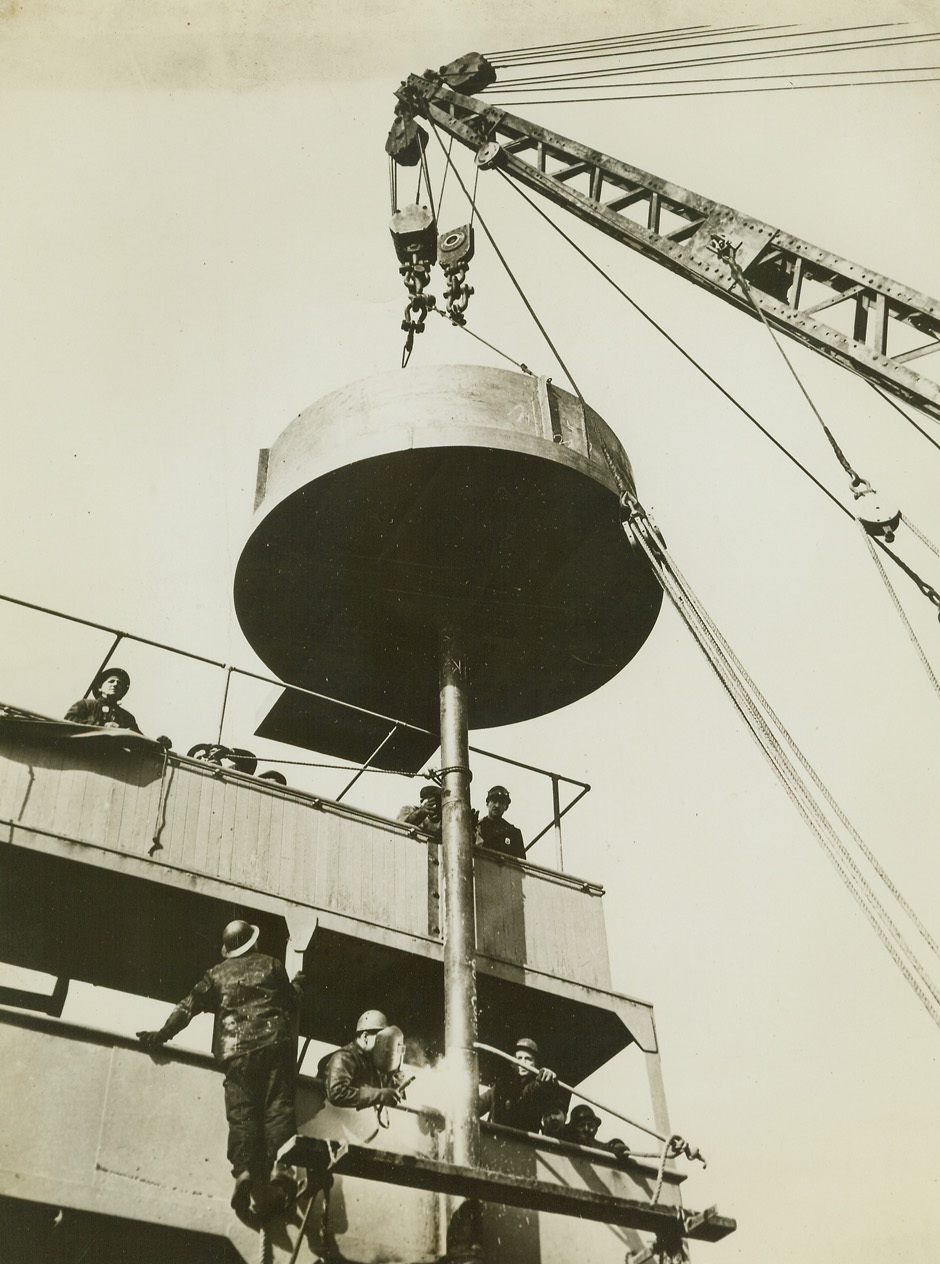 Navy Speeds Arming of Merchantmen, 4/9/1942. Held in place by a powerful crane, a machine gun nest is shown as it is welded to the superstructure of a Merchantman. Credit: Official Navy photo from ACME;