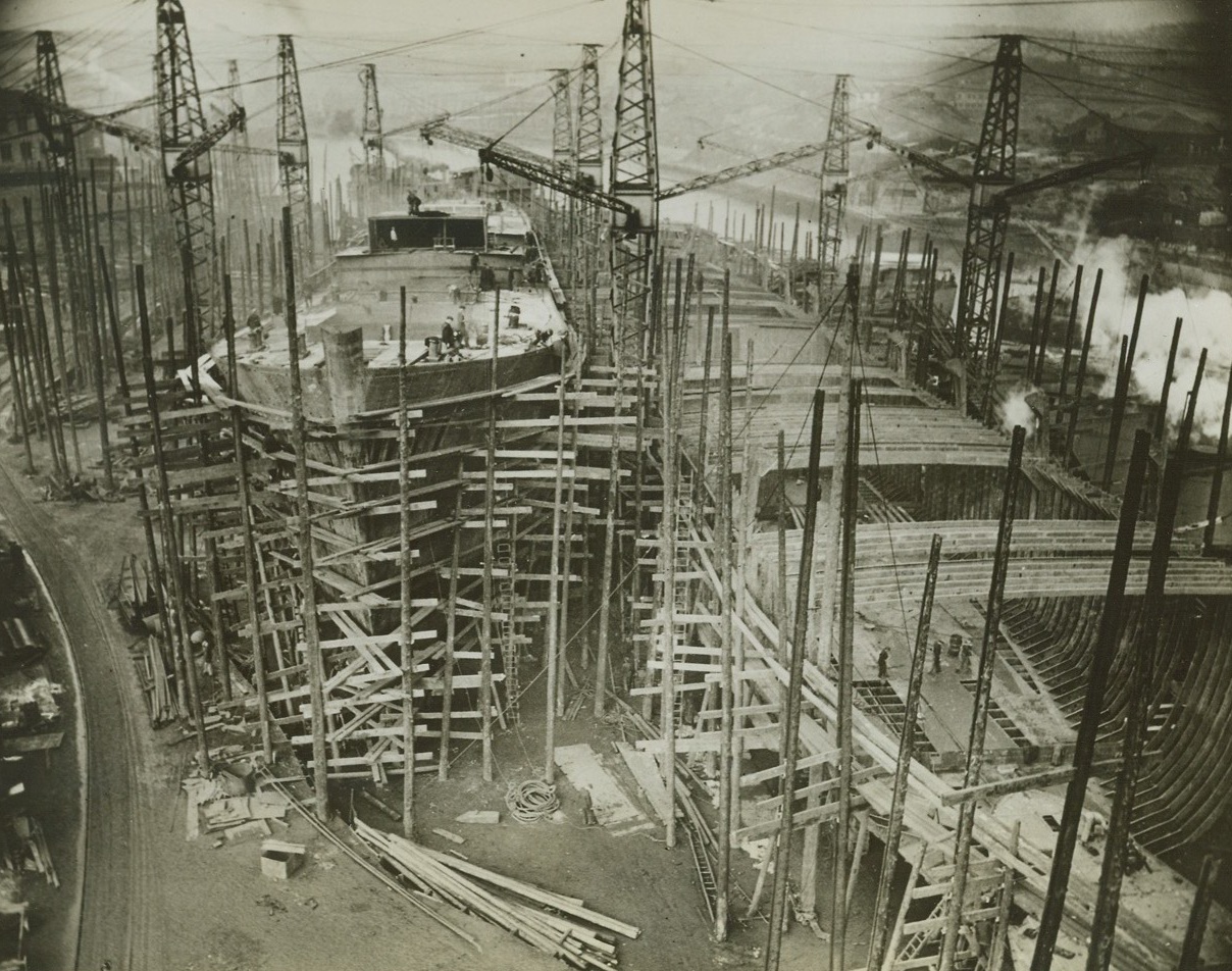 BUILDING SHIPS FOR THE BRITISH MERCHANT NAVY, 4/2/1942. AN ENGLISH PORT – British shipyards are launching ships with all possible speed; a 10,000-tower now gets its last coat of paint only 5 ½ months after the reel laying.  Widespread use of pre-fabrication has helped this speed-up and welding instead of riveting saves time and steel.  Here, is a view of two ships under construction in a shipyard at “an English port”.Credit: Acme;