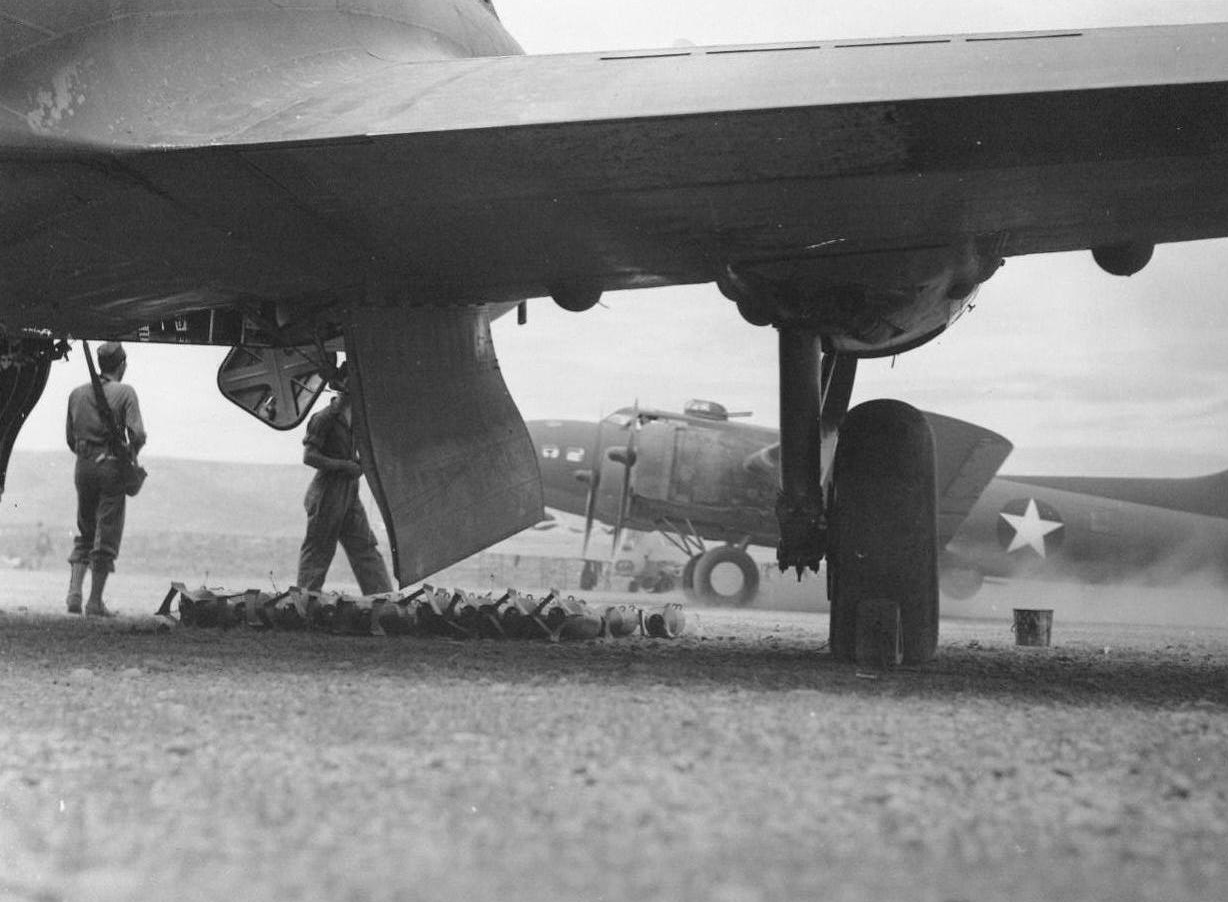 Ready To Load, 7/26/1942. In neat rows, a load of 100 pound practice bombs are ready for the bomb racks inside a B-17 flying fortress at an operational bombing base somewhere in the Northwest. Note the huge bomb bay doors swung open. In the distance a loaded plane, starting a take-off run, kicks up quite a sandstorm. To bases like this, bomber units, formed at regular Army airfields throughout the country are sent for a month’s final training before hopping off for the fighting front  7/26/42 (ACME);