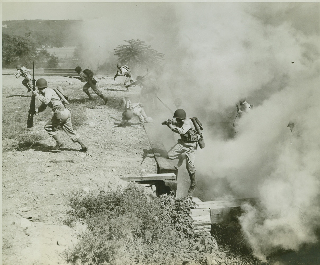 PLEBES GO OVER AND AFTER ‘EM, 8/6/1942. WEST POINT, N.Y. – West Point plebes, on a shin-barking, back-breaking “assault” course, drive through a smoke screen, hurdle a trench and tear after the “enemy”, August 6th. Carrying 22 pounds of equipment each, these boys travel a 200 yard course, hurdling 8-foot log fences, jumping wide trenches, bayoneting dummies that pop out at them unexpectedly, and meet with other obstacles with which they are not familiar. It takes a little more than four action-packed minutes to cover the course, but it teaches the boys to meet and conquer with instantaneous speed, the same sort of surprise obstacles they would meet on the field of battle. Credit: OWI Radiophoto from ACME;