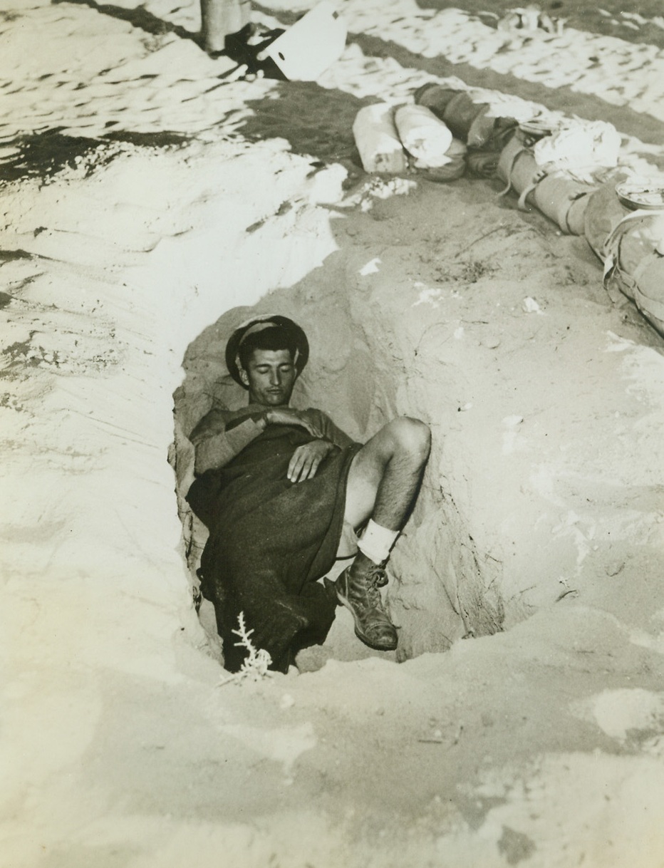 Desert Bed, 9/11/1942. Egypt – Here’s how a U.S. tankist parks himself for the night on the Egyptian desert.  He digs himself a slit-trench bed, which isn’t exactly a commodious as his bed at home but affords shelter from enemy shrapnel.  Note that the Yank, Benjamin Folsom Jr., prefers British shorts to trousers for wear under the scorching desert sun, as do many Americans in Egypt. Credit line (ACME);