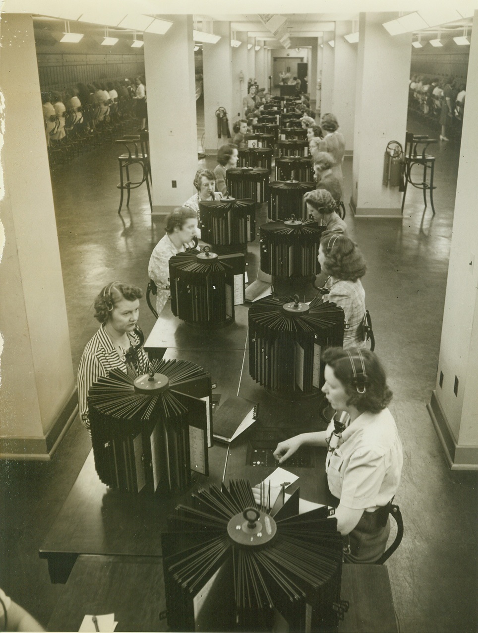 Phones of War at Work, 9/15/1942. Arlington, VA.—View of the information section in the telephone exchange of the War Department’s new office building at Arlington, just across the Potomac from Washington. It is in this section that operators look up and verify the correct branch or extension number for the many thousands of Army officers and civilians who work in the building. The exchange requires nearly 300 operators and supervisors; has more than 86,000 miles of trunk wires inside the building; has 370 incoming central trunk lines, 200 outgoing trunk lines, and 200 long distance loops. The 12 submarine canals which carry under the river to Washington weigh more than 250 tons. Credit: ACME.;