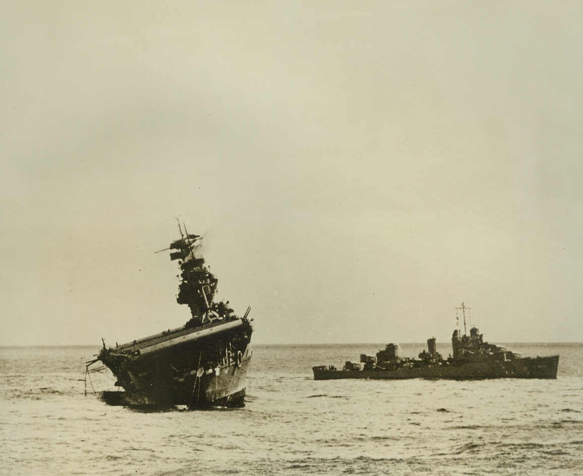 Stricken—But Still a Fighter, 9/16/1942. Washington, D.C. – An accompanying warship stands helplessly by as the stricken aircraft carrier U.S.S. Yorktown – her guns still pointed defiantly towards the sky from which Jap planes rained down bombs on her unceasingly – lists heavily toward the sea which finally claimed her.  Two torpedoes from a relentless Jap sub finished off the 19,900-ton flat-top, which sank June 7th after it had gone through the worst of the battle of Midway island, and after planes that took off from her deck exacted a heavy toll on the Jap armada headed for Midway and the Hawaiian islands.  Credit Line (Official U.S. Navy photo from ACME);