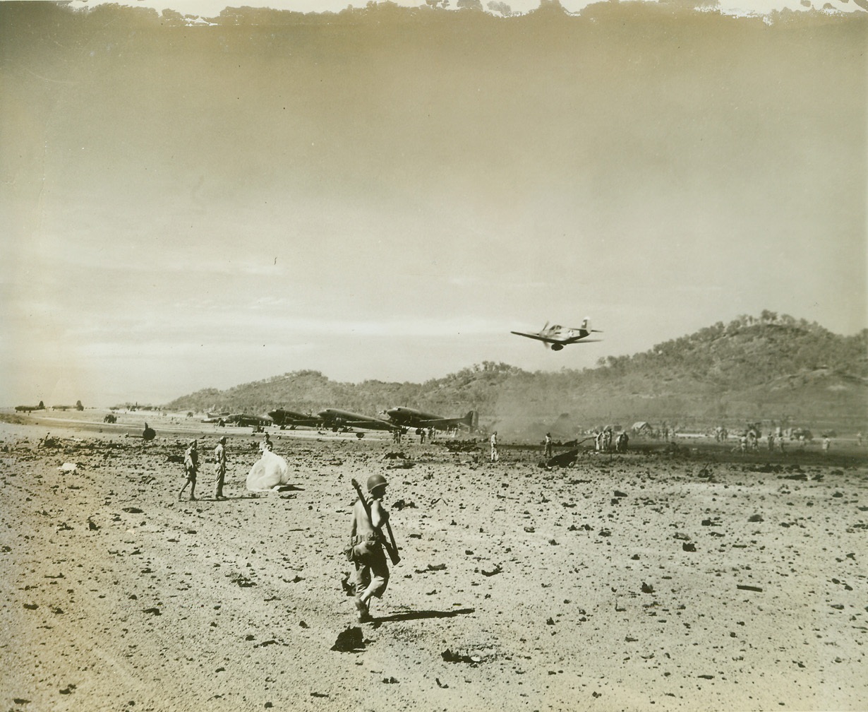A debris-strewn airfield, 9/17/1942. New Guinea—A debris-strewn airfield marks the end of a surprise Jap raid on the Allied base of Port Moresby. An American P-39 circles low over the landing field to see if the terrain is in shape for a landing. American and Australian soldiers pick up scraps of wood and metal and repair damage which was not great.  Credit: ACME;