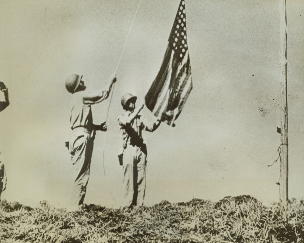 December 7—Plus Eight Months, 9/8/1942. SOLOMON ISLANDS—Marines hoist Old Glory over the first territory taken back from the Japs, eight months to the day after Pearl Harbor. Not fully showing in the picture is Major General Vandergrift, one of the leaders in the attack on the Solomons. Credit: U.S. Marine Corps photo from News of the Day supplied by ACME;