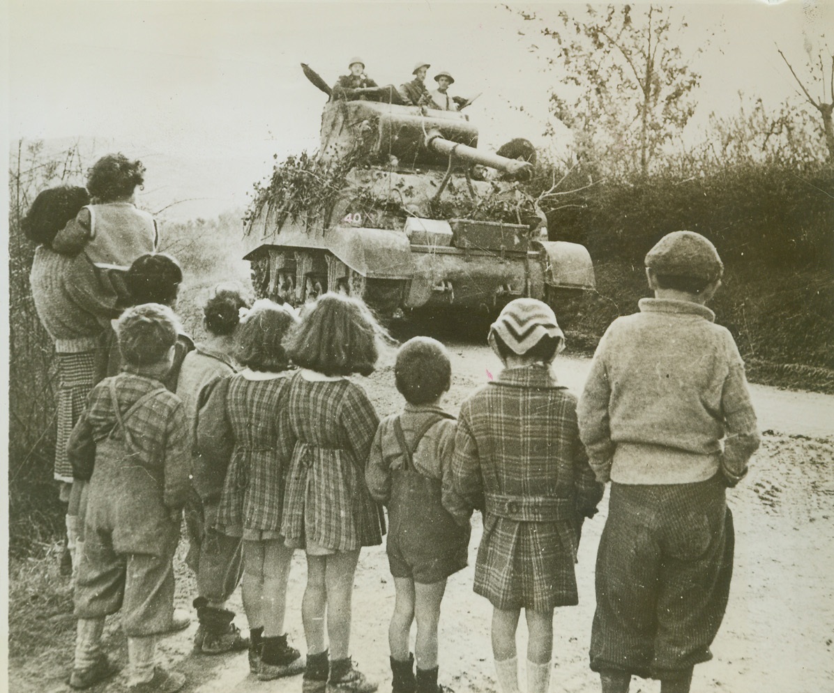 Bred to Warfare, 11/25/1944. Italy – Dressed neatly, a group of Italian children stand on the side of a road, an interested audience as a Sherman tank of the Eighth Army rumbles by. Italy’s new generation, symbolized here, has been bred to the cannon, knowing only the privations of warfare under Fascist rule. The ponderous tank, a tool of Mars, in this case is also a symbol of liberation. As the tank and its crew moves ahead the forces of Democracy and a new way of living moves in on these youngsters. Credit (British Official Photo from ACME);