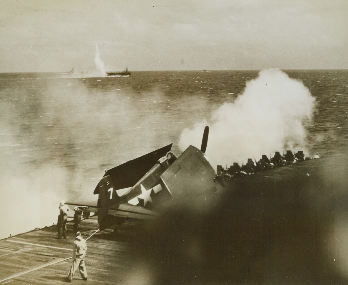 Jap Plane Kayoed by Ack-Ack, 11/17/1944. A Jap plane winds up in a watery finish after being picked out of Pacific skies by the Anti-Aircraft guns of an American Carrier. Even as the plane zooms to a splashy grave, one of its bombs scores a near-miss beside one of our carriers in the background. In the foreground, another carrier, a Grumman Hellcat spotted on the deck, peppers attacking Jap planes with salvos of shells from its anti-aircraft batteries. Exchange took place during the second battle of the Philippine Sea. Credit: U.S. Navy photo from ACME;