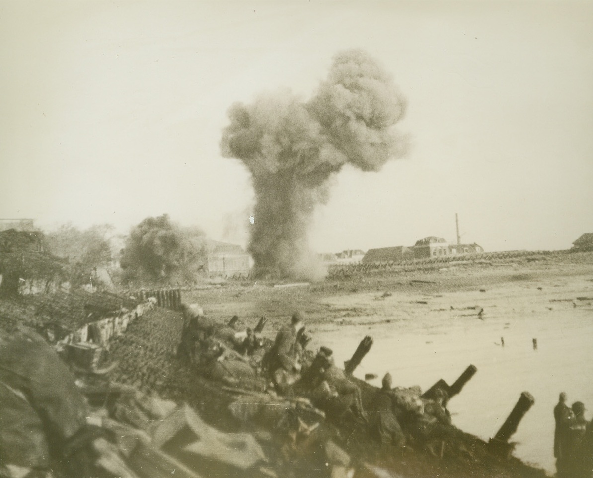 BRITISH COMMANDOS MOVE ON TO WALCHEREN, 11/10/1944. FLUSHING, WALCHEREN IS.—With geysers of earth and water rising in the background, British Commando units advance slowly along the beach at Walcheren, during the initial landings at Flushing on Nov. 1 After the first assault, the troops moved onto West Kapelle to form a second bridgehead.  Credit: BRITISH OFFICIAL PHOTO FROM ACME.;