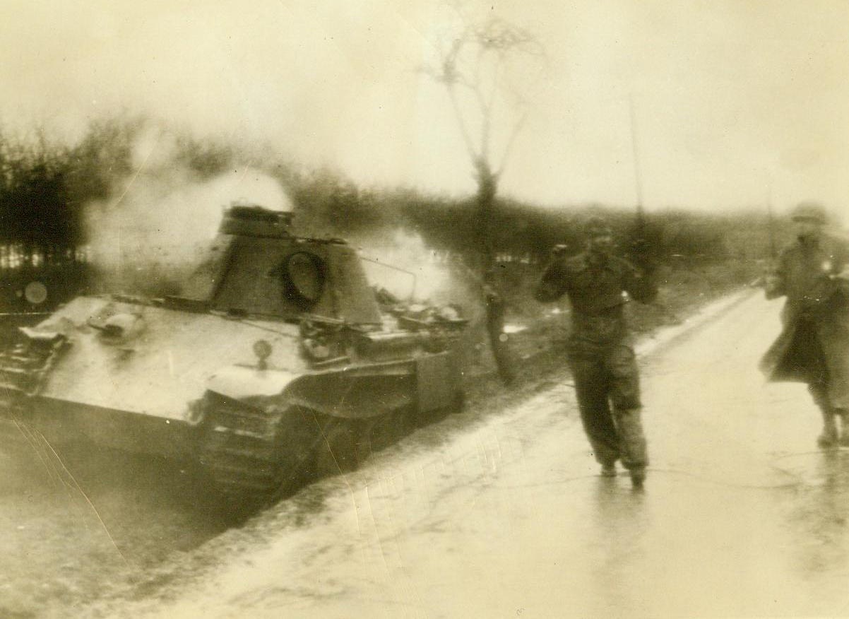 One Nazi Who Won’t Fight, 12/24/1944. Belgium – Sgt. Bernard Cook of Los Angeles, Calif., keeps his pistol pointed at a Nazi Prisoner taken as Germans attempted breakthrough in Belgium. A knocked-out German tank burns on the side of the road. 12/24/44 Credit (Army Radiotelephoto From ACME);