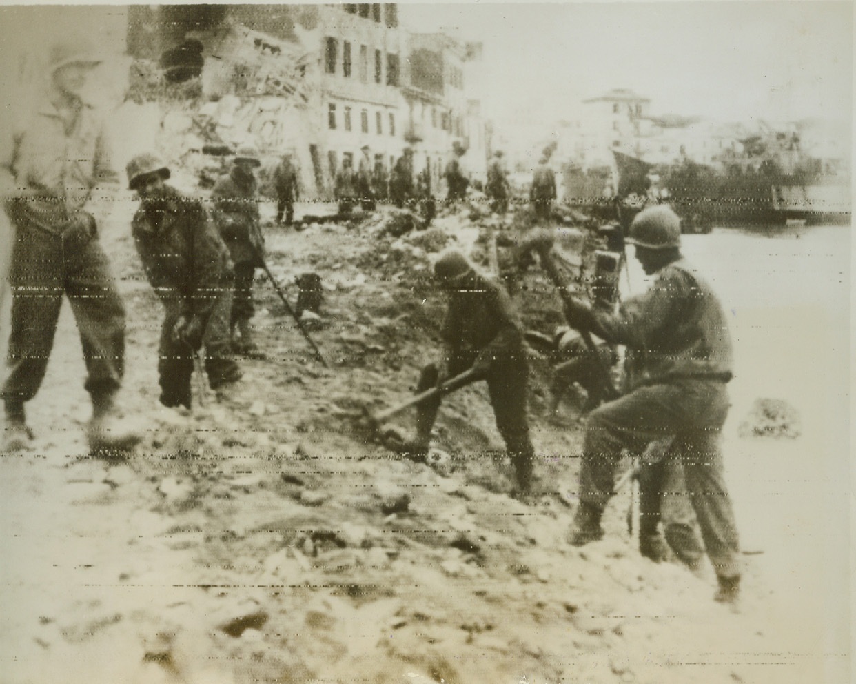 Anzio, Italy, 1/26/1944. American soldiers remove depth charges and demolition bombs planted by the Germans, along the beach at Anzio, one of the points where streams of men and material are pouring ashore for the Allied march on Rome. When Allied forces made their surprise bypass landings near Anzio, Germans tried to demolish harbor installations in the city but were not entirely successful. Note some buildings (background of photo) showing damage. Photo was flashed to New York by radio tonight.  Credit:  (Stars and Stripes photo thru OWI radiophoto from ACME);