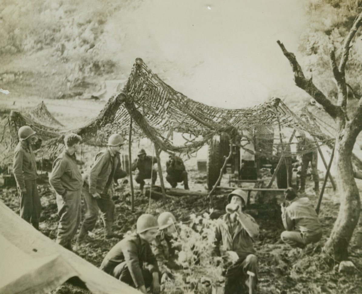 Wee Drop O’ Christmas Cheer, 1/12/1944. Somewhere in Italy—Refusing to be annoyed by the boom of big guns being fired on Nazi positions above San Vittore, members of this gun crew take turns at observing the Christmas holiday in the midst of the fight. In foreground, three Yanks gather around a make-believe Christmas tree, and one of them tilts the little brown jug for a wee bit of Yule cheer. In background, their buddies crouch and hold their ears as the gun goes off. This is an original of the radiophoto previously serviced. Credit: ACME.;