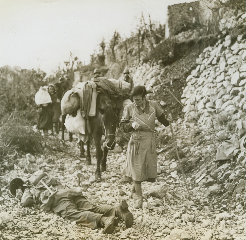 Return of the Natives, 1/1/1944. LAGONE, ITALY—Coming down from the hills in which they hid while war raged about their homes, these natives find a dead Nazi soldier in their path. Leading a mule laden with her belongings, her face distorted with hatred, an Italian woman carefully avoids fallen Nazi.  Credit: WP (ACME photo by Bert Brandt, War Pool Correspondent);