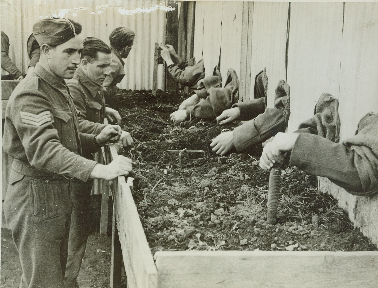 British Practice “Blind” on Enemy Mines, 1/20/1944. Italy – A device called the “Moascar Stocks” – a fence with burlap-shielded holes for arms – enables British fighters to “get the feel” of enemy mines they might have to dissemble in the dark. Instructors watch the hands of their students of the four-day school in Italy. Credit: (ACME);