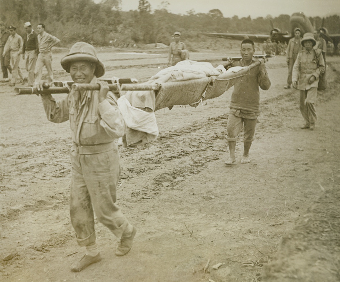 Chinese Wounded on Burma Front, 1/31/1944. Burma – Two Chinese carry a wounded companion to a dressing station on a crude stretcher made of bamboo poles “somewhere on the Burma front.” Note huge American-built transport plane in background. (Passed by censors.) Credit: ACME photo by Frank Cancellare for the War Picture Pool;