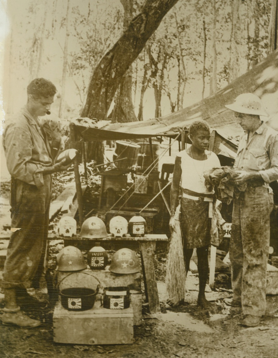 Jungle Novelty Shop, 1/6/1944. Guadalcanal – Jap soldiers unwittingly provided the “novelties” for this unique trading shop run by a Marine on Guadalcanal.  The proprietor, Cpl Robert A. Weeks (right) of Urbana, Ill., decorates the trophies he picked up on the battlefields with Jap flags, Jap characters, etc.  He is shown bargaining for a souvenir with a native boy as Cpl Joseph F. Andrejka, Chicago, Ill., examines a decorated Jap canteen.  Barely discernible on Cpl Andrejka’s shoulder is “Coco”, a variety of possum which is found on the island and is very easily tamed.;