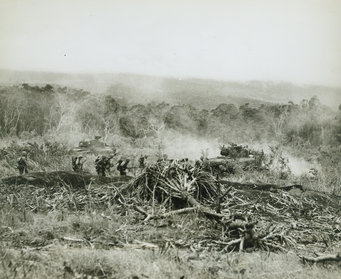 Tanks Lead the Way, 1/18/1944. New Britain – With their tanks spitting fire as they lead our fighters into the babble for Cape Gloucester airfield, hunched Leathernecks follow close behind to take care of all Jap snipers who might be in nearby trees. Battle smoke begins to cloud the fighting field as the boys creep past a large bomb crater made by our raiders before the big push. Credit line –WP- (ACME photo by Frank Prist, Jr for the War Picture Pool;