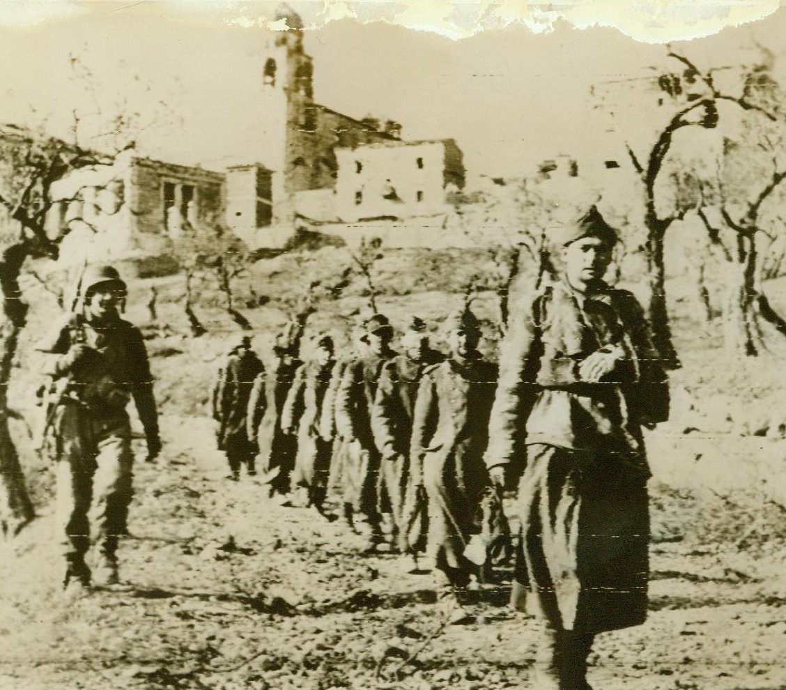 War-Weary Men in a War-Torn Village, 1/11/1944. Italy— Captured Germans are marched away from crumbling San Vittore, Italian town blasted by Nazis and Americans. The enemy soldier leading the line wears a sling, while another casualty follows in file. 1/11/44 ACME;