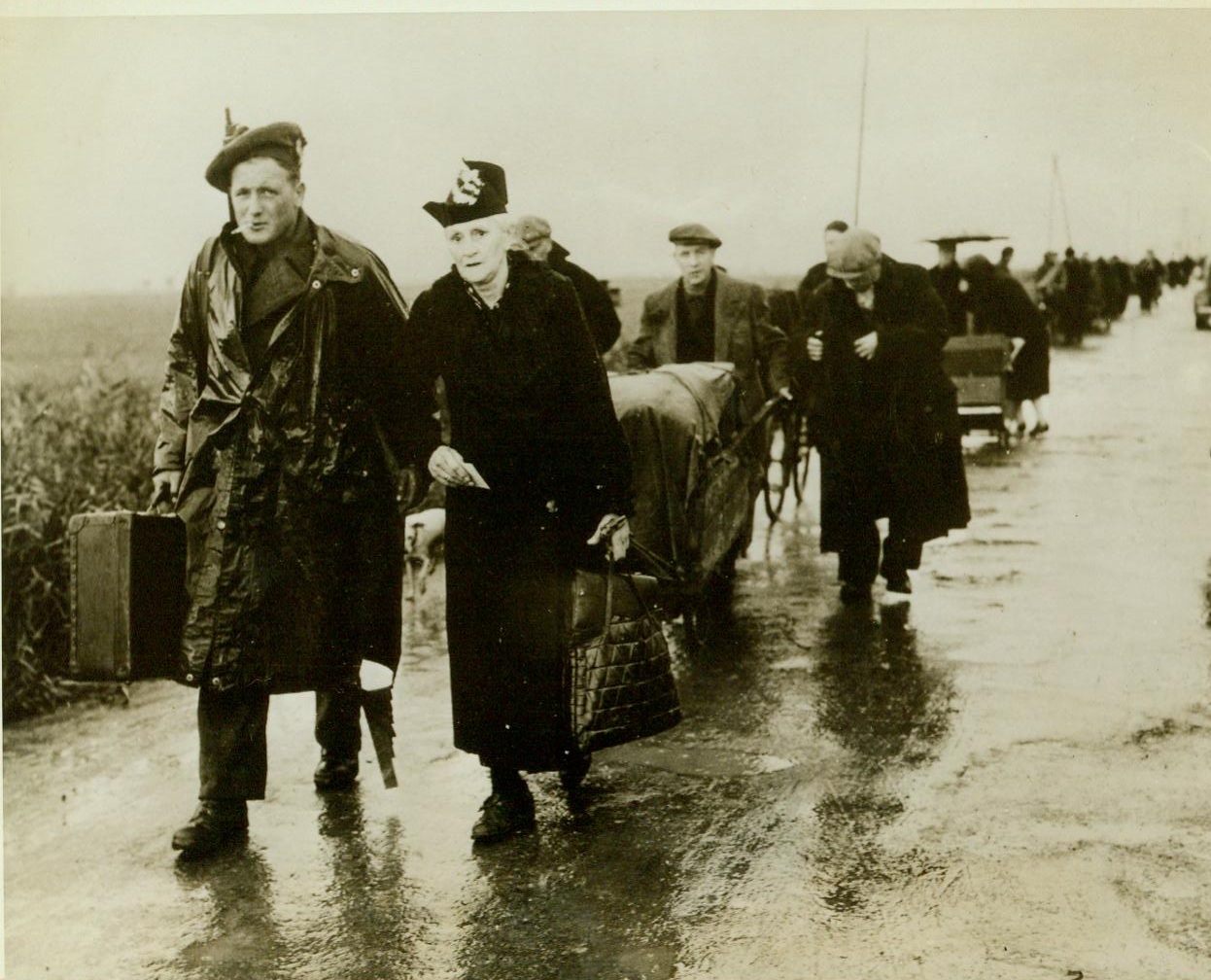 Exit from Dunkirk, 10/9/1944. Dunkirk, France – Walking beside a British Soldier who carries her bag, a white-haired woman of Dunkirk leaves the besieged city during the 60-hour truce. Behind her, a long column of civilian refugees trudges the rain-soaked road away from Dunkirk. Credit: –WP-(ACME);