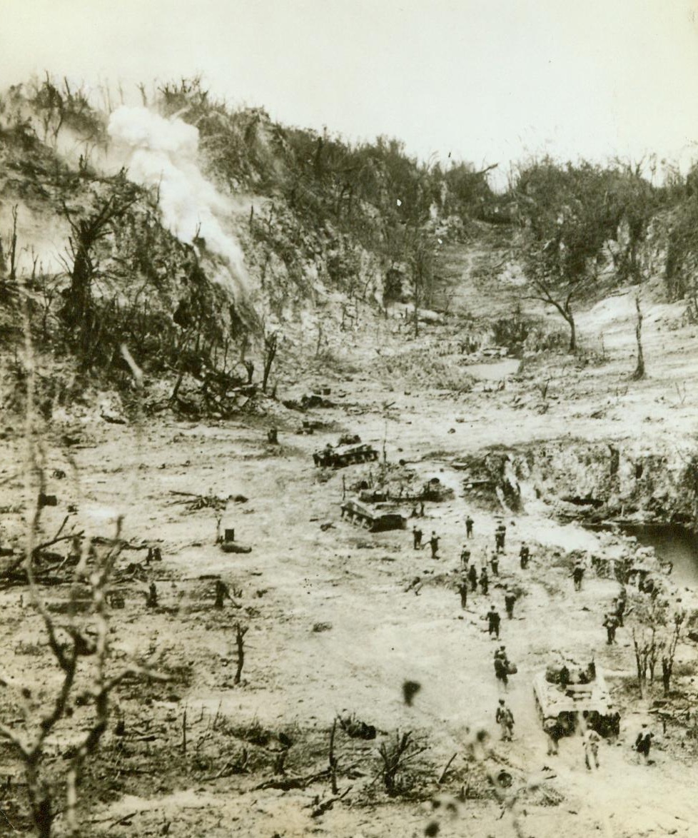 Mop Up On Peleliu, 10/18/1944. Marine Infantrymen, supported by tanks, move up to attack enemy nests in a hillside on Peleliu. The smoke at left marks the blast of a shell fired by one of the tanks. The do-or-die remnants of the Jap forces on the island had to be cleaned out of strong positions in caves and dugouts in the many ridges on Peleliu 10/18/44 Credit (Marine Corps Photo From ACME);