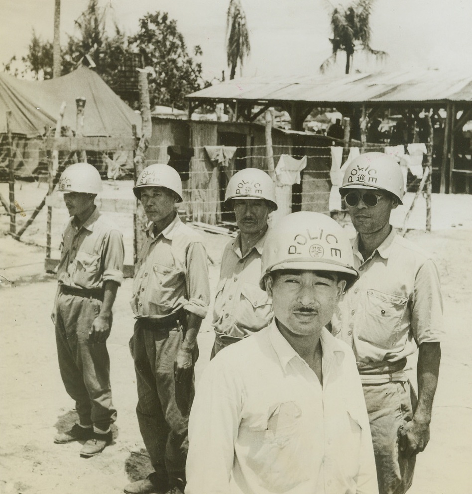 Civilian Life on Saipan, 10/27/1944. Saipan, Marinas – To Hirasawa, former civic official of Aslito, (foreground) goes the job of chief of Japanese Internal Police on Saipan.  He chose his own men, who are very proud of their new helmets, denoting their authority.  Here they stand in front of the “hans”, or huts in the civilian camp.  The shelters were made of tin sheeting from the bombed sugar refinery and weathered planks.  Each one houses from 20 to 55 people.Credit line (ACME) (WP);