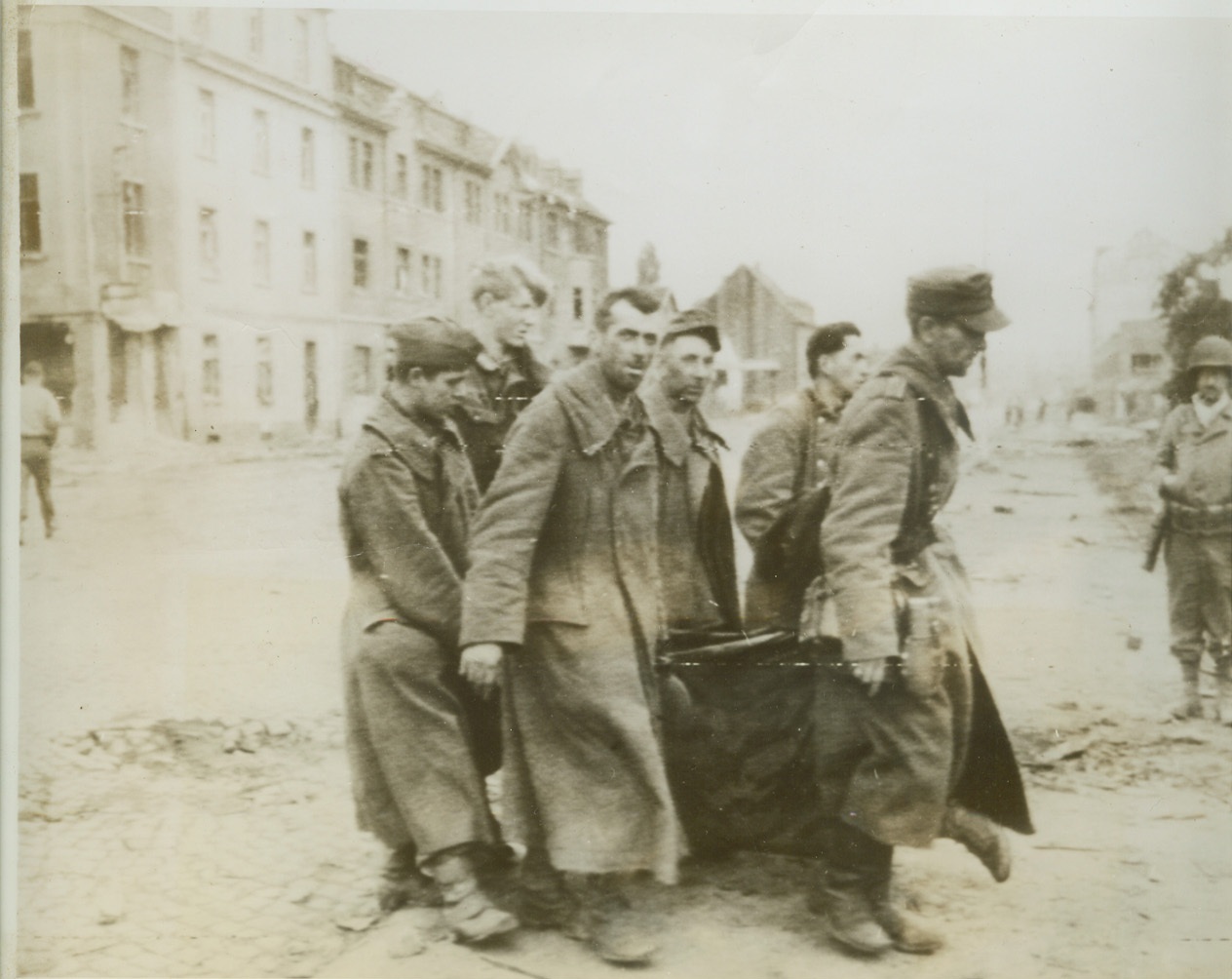 Aachen Prisoners Remove Own Wounded, 10/14/1944. GERMANY – Haggard and worn, this group of Nazi prisoners, taken during the battle for Aachen, uses a tent to remove a wounded comrade to an aid station. An American soldier stands aside watching the captured Germans. Credit (Army Radiotelephoto from ACME);