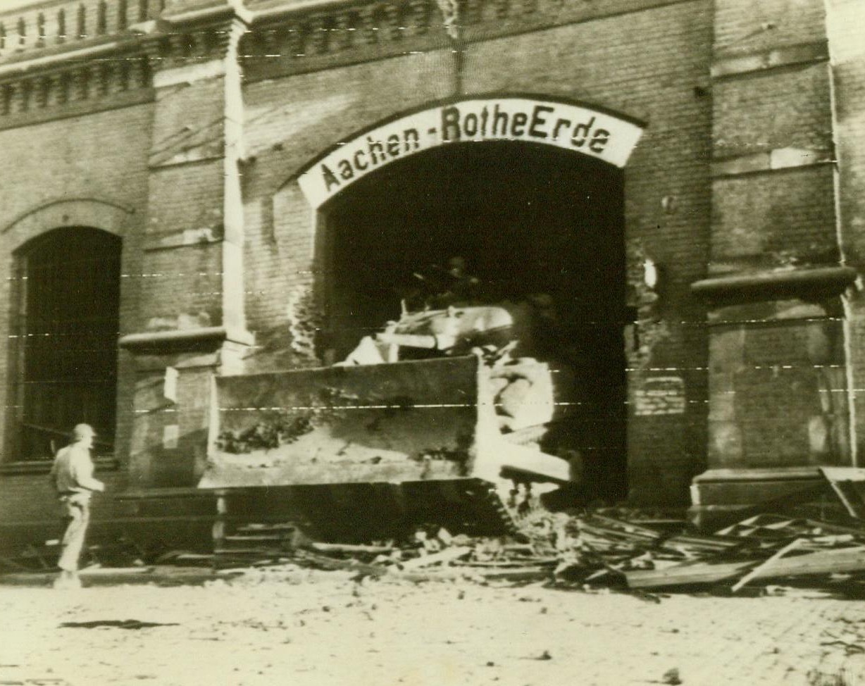 Smashing Way Into Aachen, 10/17/1944. Germany – An American tank, with bulldozer attachment, smashes its way through a hole in the Aachen Railway Station to enter the city proper after German garrison had blown up the main viaduct. 10/17/44 (ACME);