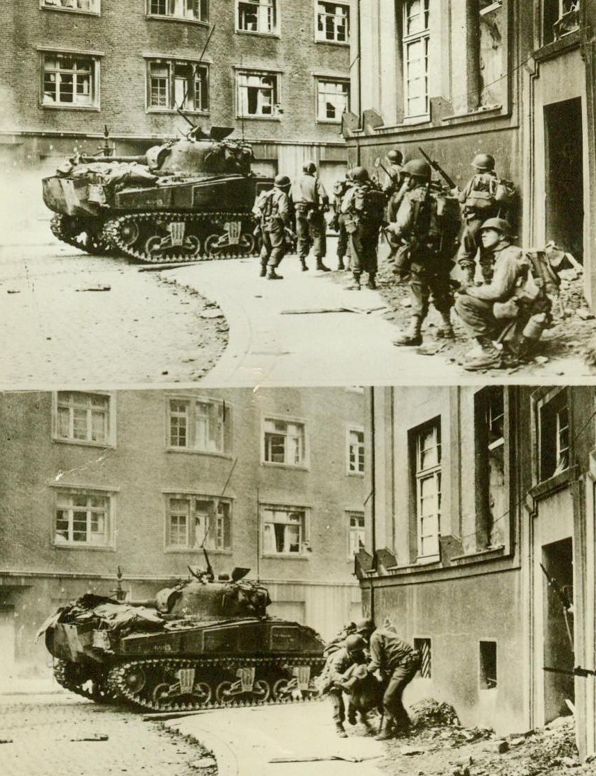 Yanks Move Up In Aachen Battleground, 10/20/1944. Aachen, Germany – As the fight for Aachen, gateway to the Rhine, continues, American doughboys move forward in street-to-street fighting. In the upper photo, an American tank and a group of infantrymen inch slowly forward, ever on the alert for German snipers hidden in the buildings. In the lower photo, as the rest of their group moves forward, two Yanks carry a wounded buddy back to the safety of a building already cleared out. American forces are officially reported to be in possession of more than half of the city, including the main railroad station. 10/20/44 (ACME);