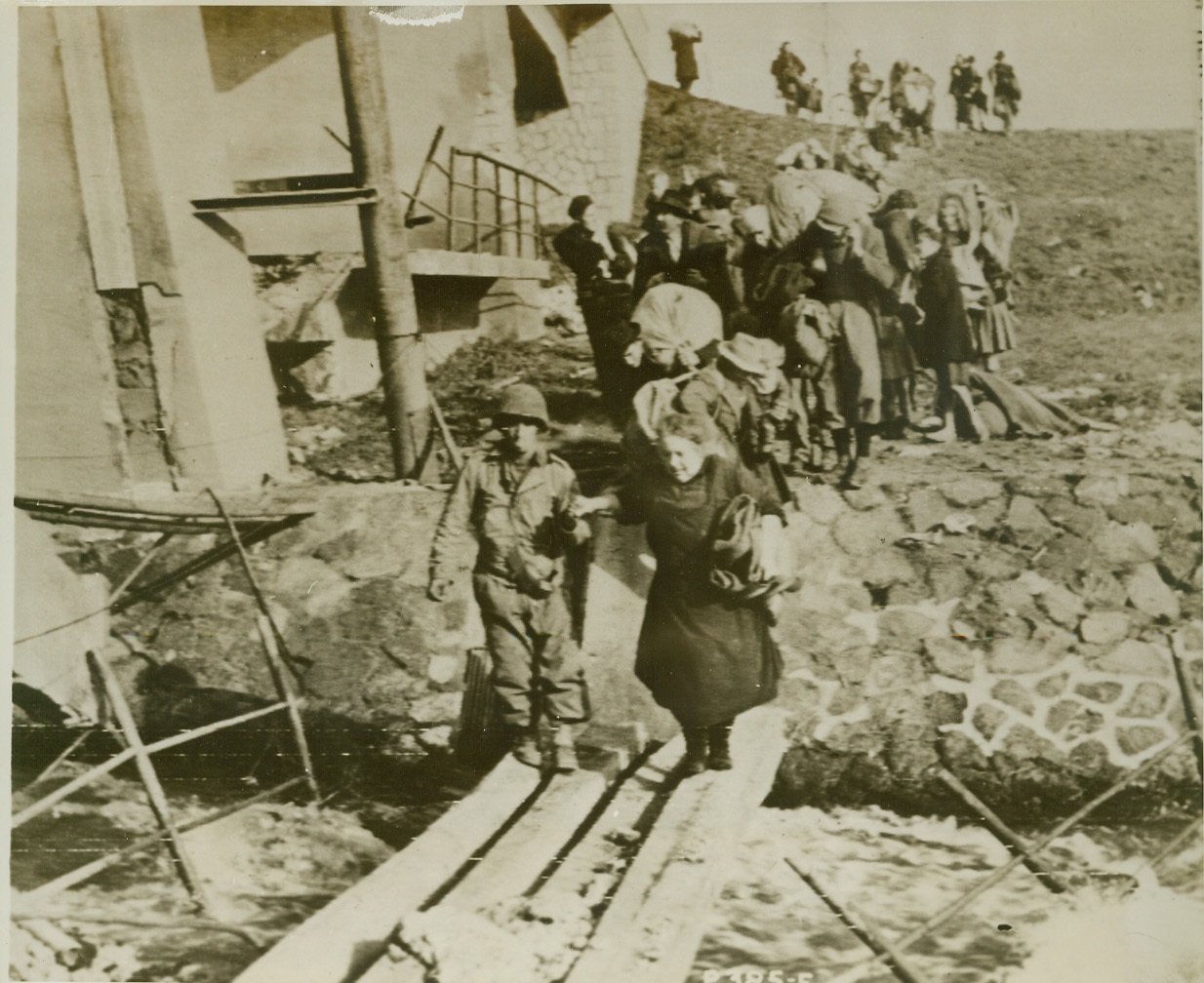 Yanks Lend a Hand, 2/4/1944. Italy – Pfc Manuel Martins of New Jersey lends a helping hand to an aged Italian woman crossing a makeshift bridge in the Mussolini Canal Sector of Italy. Behind them are other refugees, making their way back to their homes in territory now occupied by the Yanks. Credit: (ACME photo by Charles Seawood for the War Picture Pool, transmitted via U.S. Army Signal Corps Radiotelephoto);