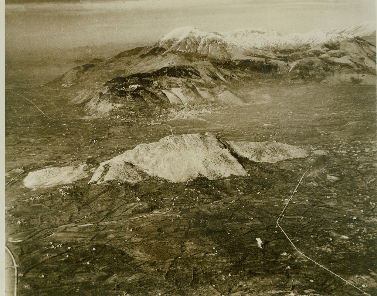 War-Torn Valley on Road to Rome, 2/6/1944. Italy – A photo from an Allied observation plane, looking north over the Cassino Battlefield, gives a bird’s-eye view of the embattled valley on the road to Rome. The burst of smoke in the right foreground reveals an American gun position firing on Nazi installations in Cassino, the shell-torn town huddled at the foot of the mountain ridge in the background. With the capture of Mt. Trocchio (center) and Mt. Cairo, high peak in the background, Fifth Army fighters have three-quarters circled the enemy, the Abbey of Monte Cassino, perched in the mountain above Cassino (left of town), bristles with Nazi guns trying to thwart our drive to Rome. The Rapido River is at top right. Credit: (ACME);