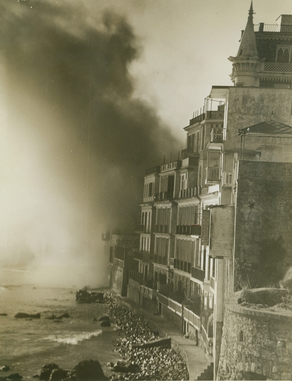 Nettuno Waterfront Under Attack, 2/26/1944. Nettuno, Italy – Most of them obscured by heavy shell smoke, the beautiful homes lining the waterfront at Nettuno are targets for the Nazis. Pounding the Allied holdings from their positions along the Italian coastline north of Anzio, the Germans keep up a relentless barrage. Credit-WP-(ACME Photo by Bert Brandt for the War Picture Pool);
