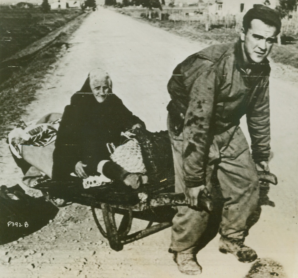 HAND-TRUCKING THE AGED TO SAFETY, 2/7/1944. ANZIO, ITALY—Pvt. Joe Maniscalo, of Brooklyn, New York, believes in chivalry as he wheels an aged Italian woman in a native hand-truck on the road to Anzio. The aged woman fled with her meager personal effects from the German-held territory near Cisterno.Credit: Official Signal Corps radiotelephoto from Acme;