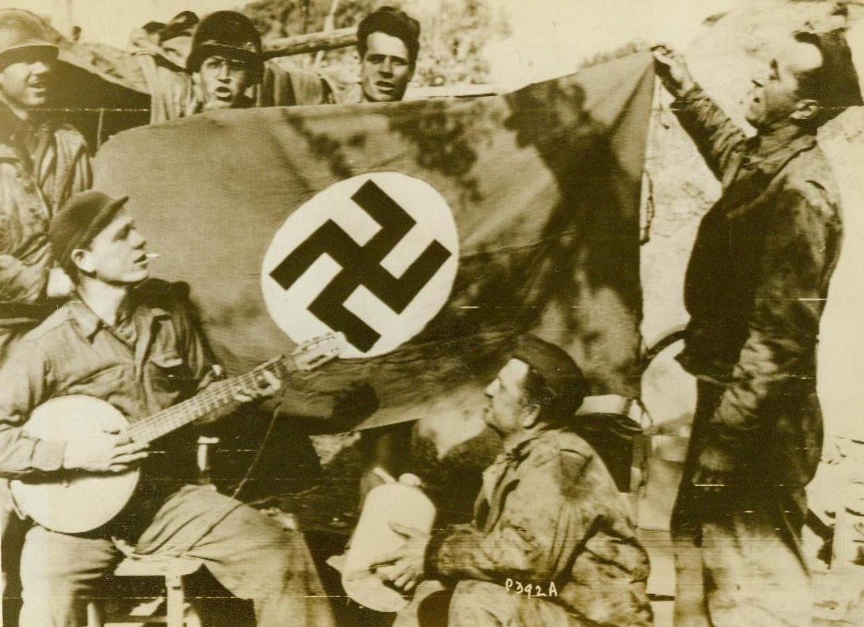 SERENADING DEFEATED ENEMY, 2/7/1944.SOUTH OF ROME, ITALY: Six Yanks break into improvised song about the Nazi Swastika flag that they captured from the enemy near Nettuno, the men, left to right, are : Pfc. Harry C. Belle, of Kingsport, Tenn; Pfc. John Gursky, of Philadelphia, Pa., an Pfc. Peter Safrey, of New York City; rear: Corp. John Cugnini, of Durango, Colo; Pfc. Wilbur Morirarity, of Lancaster, Calif; and Pvt. Kenneth B. Lance, of Toly, Texas Credit (Official Signal Corps Radiotelephoto From ACME);