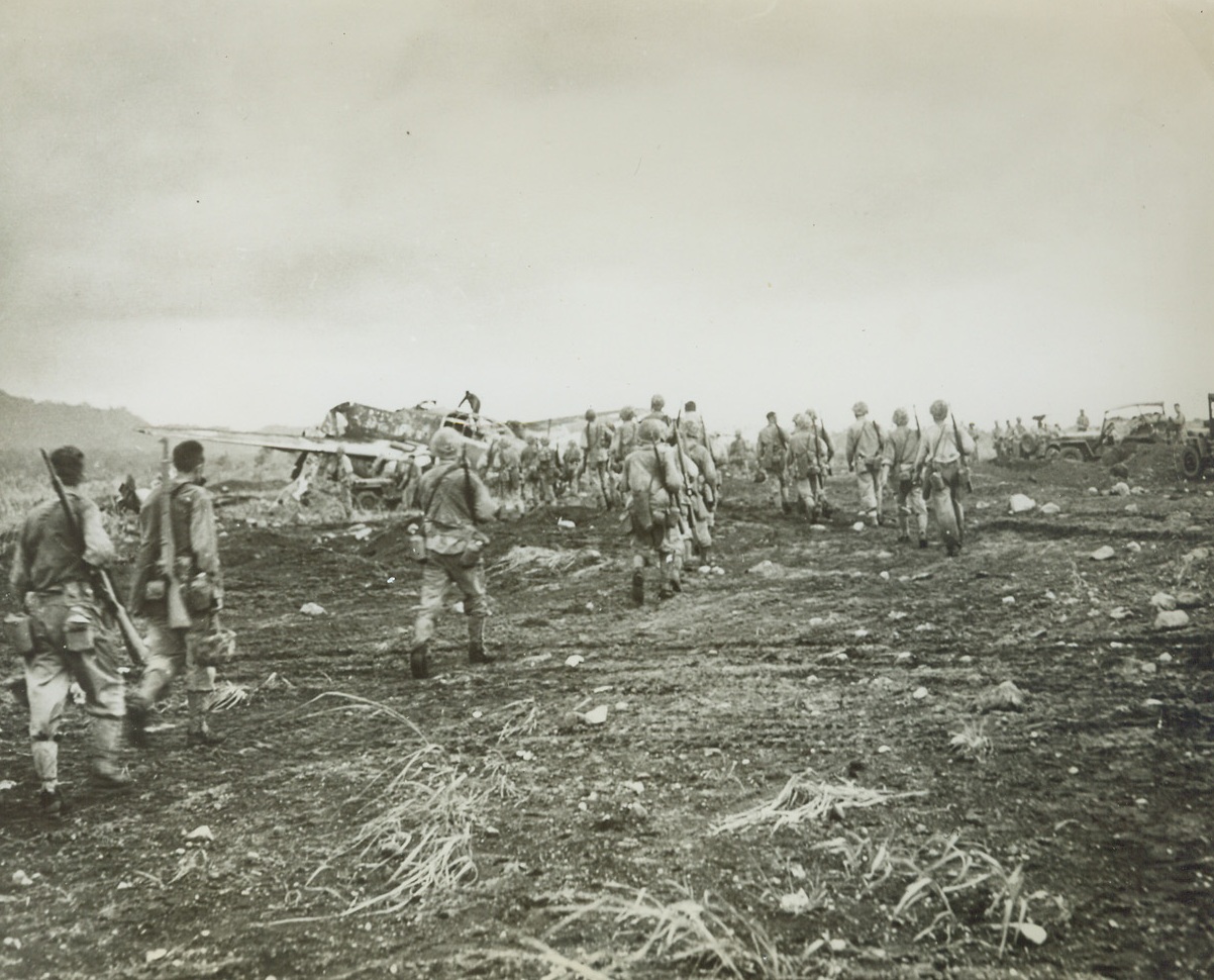 Their Objective Reached, 2/3/1944. Cape Gloucester, New Britain – After three days of hard fighting in the jungles of New Britain, Uncle Sam’s marines reach their Cape Gloucester objective—the strategic Jap-held airfield. At left, background, is a wrecked Jap plane, ruined during the heavy bombardment that preceded the leatherneck invasion. Credit: U.S. Marine Corps photo from ACME;