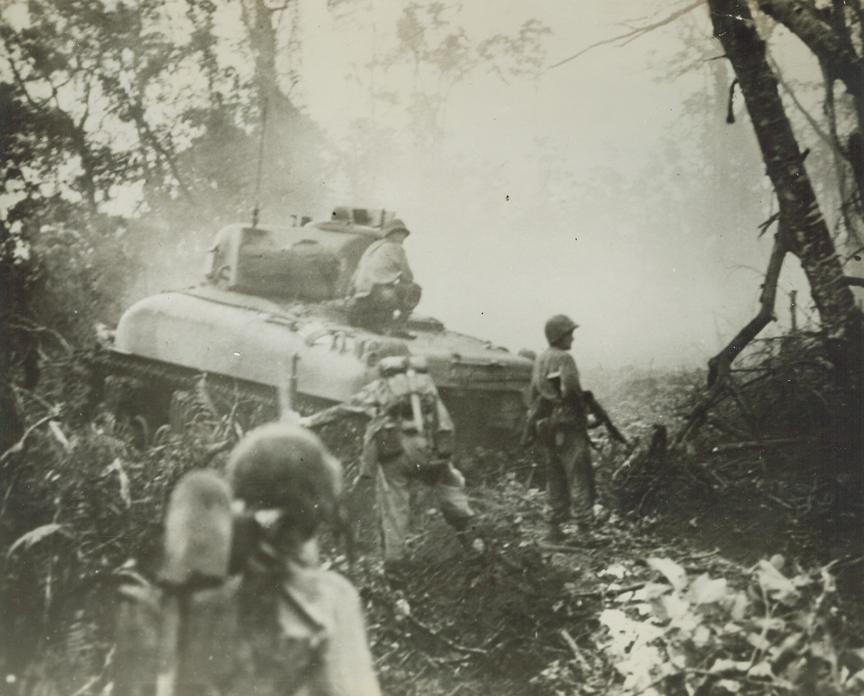 En Route to the Airfield, 2/3/1944. New Britain – Marine riflemen keep a sharp lookout for snipers and attacking enemy warriors as one of their tanks pauses on New Britain to blast away at a Jap pillbox with 75mm and machine fun fire. The leathernecks are en route to Cape Gloucester airfield, which fell to them shortly after this photo was made. Credit: U.S. Marine Corps photo from ACME;