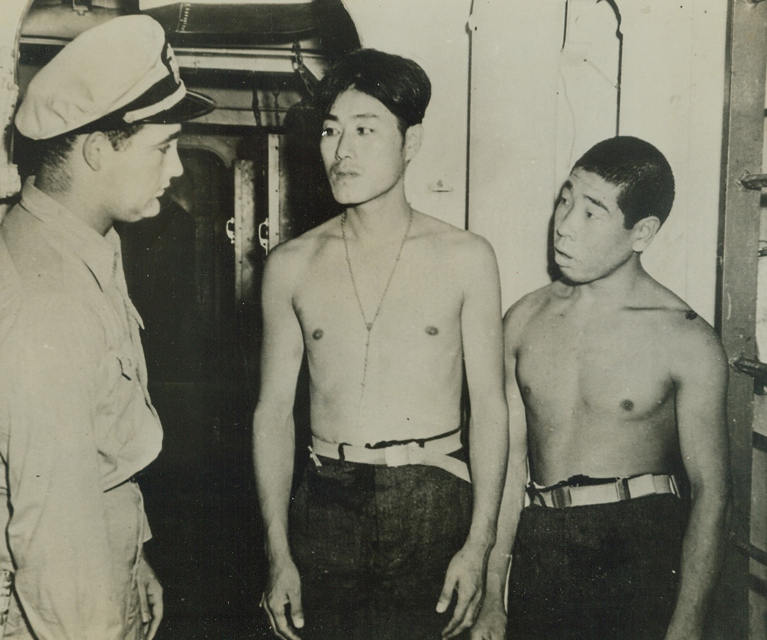 Jap Prisoners Seized in Gilberts, 2/1/1944. South Pacific – An American Naval officer questions two prisoners seized in a raid on an atoll in the Gilbert Islands. The taller of the two, wearing a rosary, claimed to be a Korean—one of the thousands of his countrymen conscripted by the Japs for work battalions. A Jap at the right at first claimed to be a laborer, too, but the Korean put him “on the spot” by signaling to the Naval officer by waving his arms that the man was a Jap flier. Credit: U.S. Army Air Forces photo from ACME;