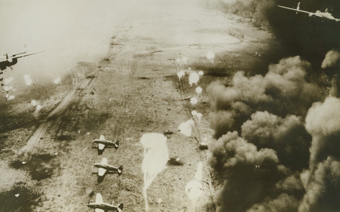 Yanks Raid Jap Base at Dagua, 2/19/1944. Dagua, New Guinea - - Parachute bombs drift down toward Jap Zeros parked on Dagua airfield and smoke rises from the besieged area as B-25 Mitchell bombers of the Fifth Air force stage a surprise raid on the enemy base.  The smoke is coming from an oil dump fire which was started by an earlier wave of bombers.  The Japs were caught completely unaware.Credit (U.S. Army Air Forces photo from ACME);