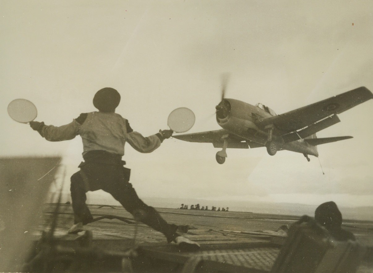HELLCAT FLIES FOR THE BRITISH, 2/10/1944. AT SEA—An American-built Grumman Hellcat fighter of the British Fleet Air Arm, comes in for a landing aboard the escort aircraft carrier H.M.S. Ravager “somewhere at sea”, as a signalman gives the all clear.  Credit Line (ACME);