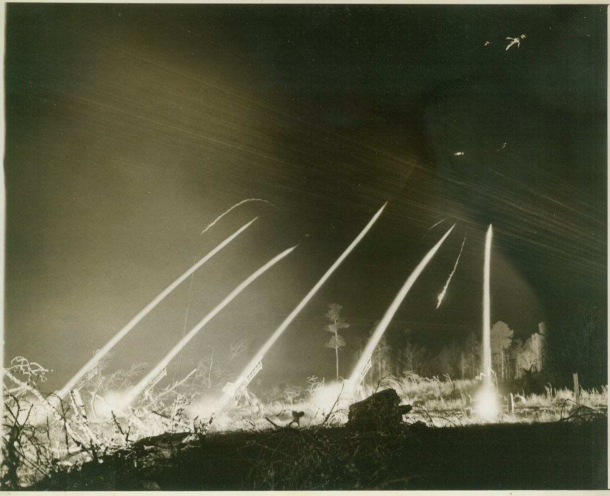 ROCKET TARGETS FOR ACK-ACK TRAINING, 3/8/1944. CAMP DAVIS, N.C. – Jet-propelled rocket targets are now being used to train anti-aircraft artillerymen at Camp Davis, giving our gunners practical experience in firing at “enemy planes” diving at terrific speed, and preparing them for action against the jet-propelled planes of tomorrow, should the enemy perfect such planes before the war is over. Fired from specially designed carriages, the rockets have an initial velocity of 450 miles per hour or 675 feet per second. They are released from the range at Holly Shelter, a Camp Davis firing point. Manning 50 caliber machine guns and 20 and 40mm weapons, the trainees improve their aim and accuracy as they track the fast-moving, 59-inch targets. The rocket always describes an approximate parabola and has a maximum horizontal range of about 2200 yards. The following photos show phases of rocket target practice by night and by day. NEW YORK BUREAU Streaking upward in thick, white streams, the rockets have barely left their launching carriages before they are caught in a web of anti-aircraft fire. Some of the jet-propelled targets have already been nipped, and their remains fall in thin streaks toward the ground. Credit: ACME;