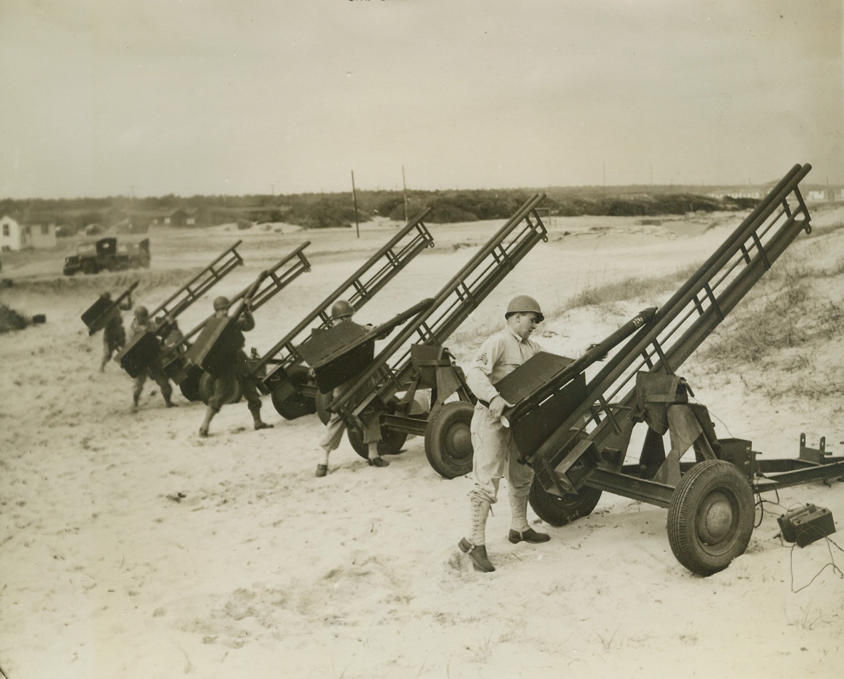 ROCKET TARGETS FOR ACK-ACK TRAINING, 3/8/1944. CAMP DAVIS, N.C. – Jet-propelled rocket targets are now being used to train anti-aircraft artillerymen at Camp Davis, giving our gunners practical experience in firing at “enemy planes” diving at terrific speed, and preparing them for action against the jet-propelled planes of tomorrow, should the enemy perfect such planes before the war is over. Fired from specially designed carriages, the rockets have an initial velocity of 450 miles per hour or 675 feet per second. They are released from the range at Holly Shelter, a Camp Davis firing point. Manning 50 caliber machine guns and 20 and 40mm weapons, the trainees improve their aim an accuracy as they track the fast-moving, 59-inch targets. The rocket always describes an approximate parabola and has a maximum horizontal range of about 2200 yards. The following photos show phases of rocket target practice by night and by day. NEW YORK BUREAU One man is all that’s needed to load one of the speedy jet-propelled targets onto the strange-looking launching carriage. One fin fits into the narrow groove between the railings that form the nose of the carriage. Here, five of the launchers are loaded at the same time. Credit: ACME;