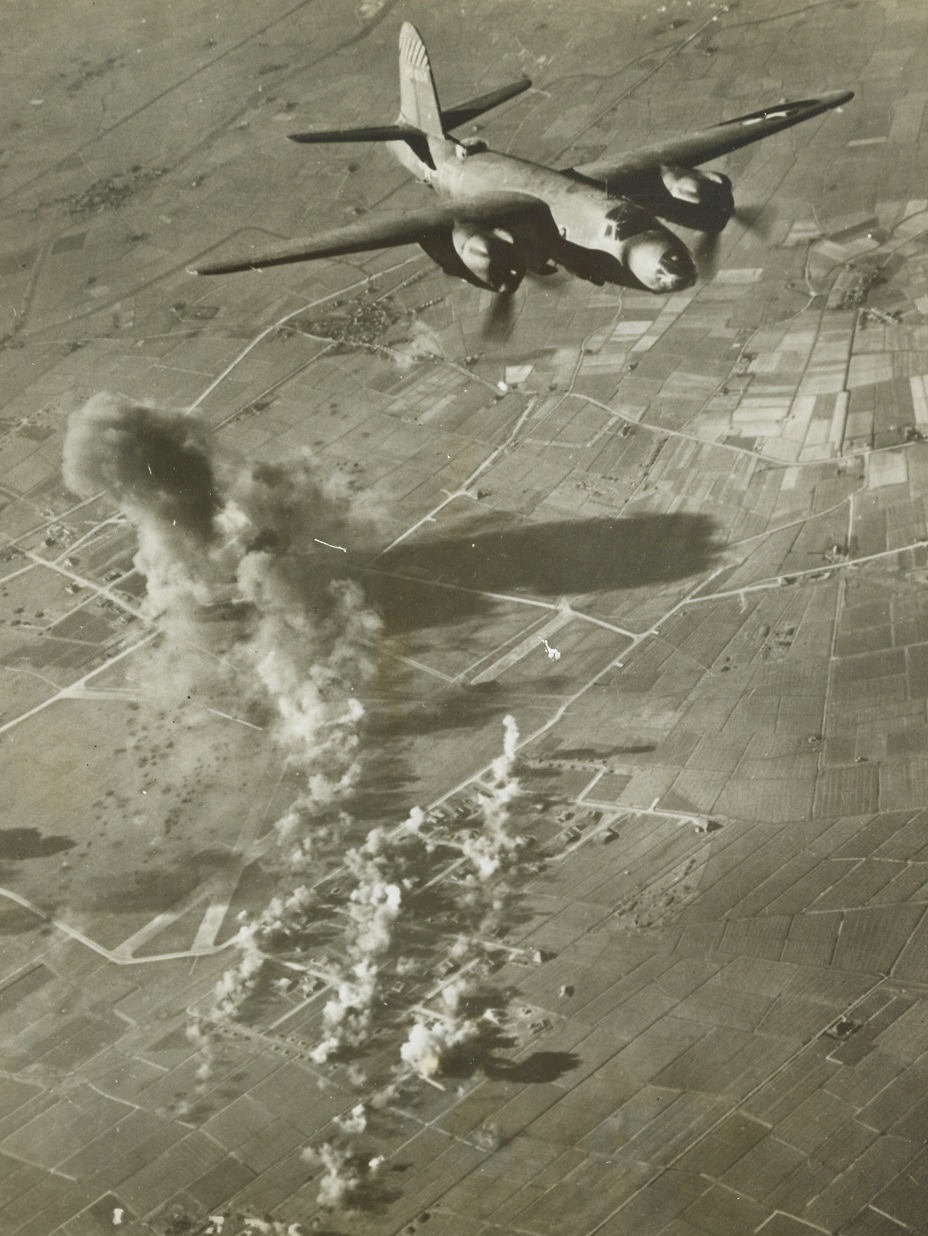 NAZI AIRDROME GETS A SCIENTIFIC MAULING, 3/9/1944. LEEUWARDEN, HOLLAND—Her bomb-bay doors wide open, a B-26 marauder roars over a Nazi airdrome at Leeuwarden, Holland where three paths of bombs cut squarely through a large dispersal area containing numerous aircraft shelters. A concentration of bombs has hit a fuel dump at the other end of the German base while at the extreme left a shadow cast by smoke rises from still another dispersal area (not shown in photo). The Ninth Air force medium bombers are continuing to give enemy airdromes in western Europe a scientific pounding.Credit: Acme;