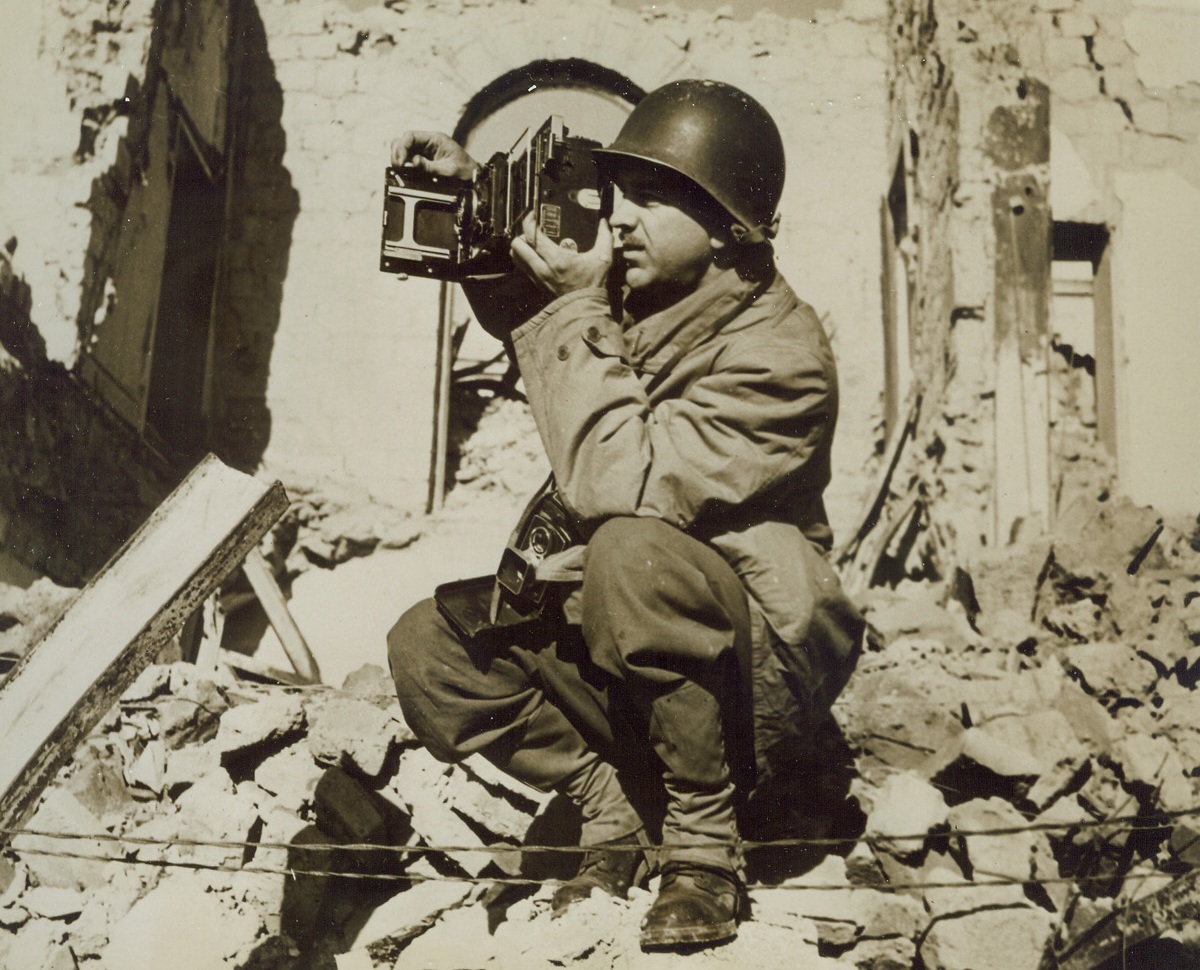 Get That Picture!, 3/3/1944. NETTUNO, ITALY – Squatting on a pile of bomb-debris, Charlie Seawood, Acme Newspictures photographer for the war picture pool, is ready to make a picture of the bomb ruins of battle-scarred Nettuno. Credit Line – WP – (Acme)A;