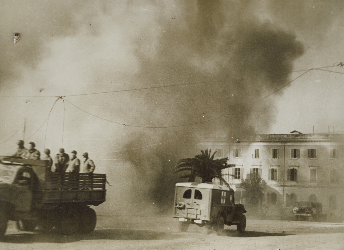 Nettuno Shelled, 3/29/1944. NETTUNO, ITALY -- An American Field Service ambulance and a truck carrying soldiers pass through the dust expelled from a 88mm shell, which exploded in the main square at Nettuno, Italy. Credit Line (ACME);