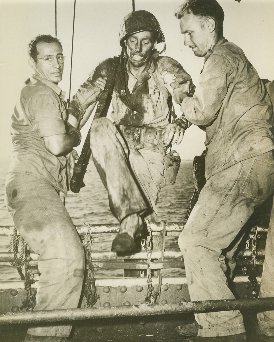 Back from Eniwetok “Hell”, 3/9/1944. Washington, D.C. – Coast Guardsmen assist a battle-blackened Marine over the side of an assault transport after two days of intensive fighting had wiped out the Japs at Eniwetok Atoll in the Marshalls.  That’s not camouflage on his face, but coral dirt which smeared his face as he burrowed into the ground to avoid being hit by Nip snipers. Credit (U.S. Coast Guard photo from ACME);