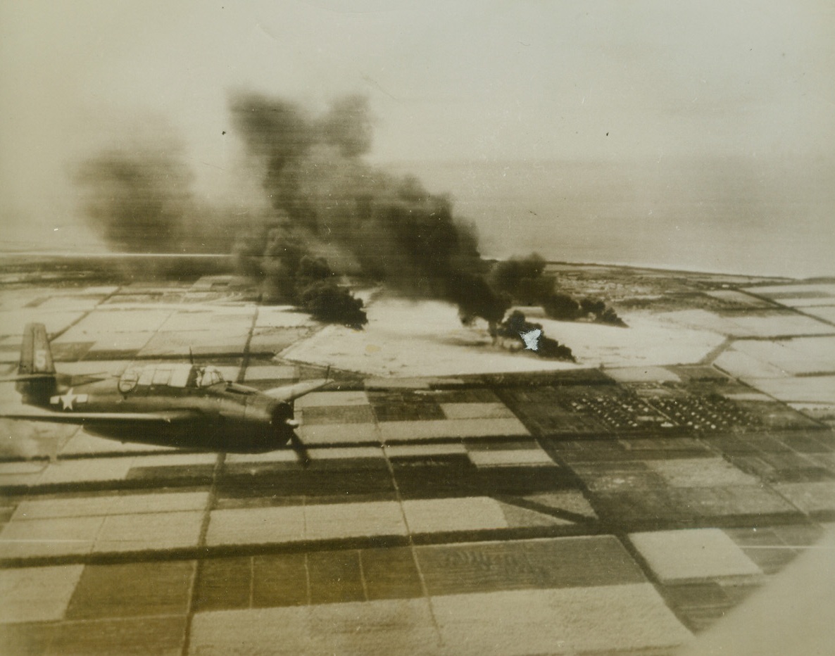 Navy Planes Blast Mariana Island, 3/7/1944. Tinian Island – Huge pillars of smoke rise from direct hits on Jap planes and supplies scored by Navy carrier-based dive bombers during a raid on Tinian island in the Marianas.  One of the planes which blasted the Japs (left) soars above the orderly squares of the sugar cane fields adjoining the enemy airstrip. Credit line (ACME);