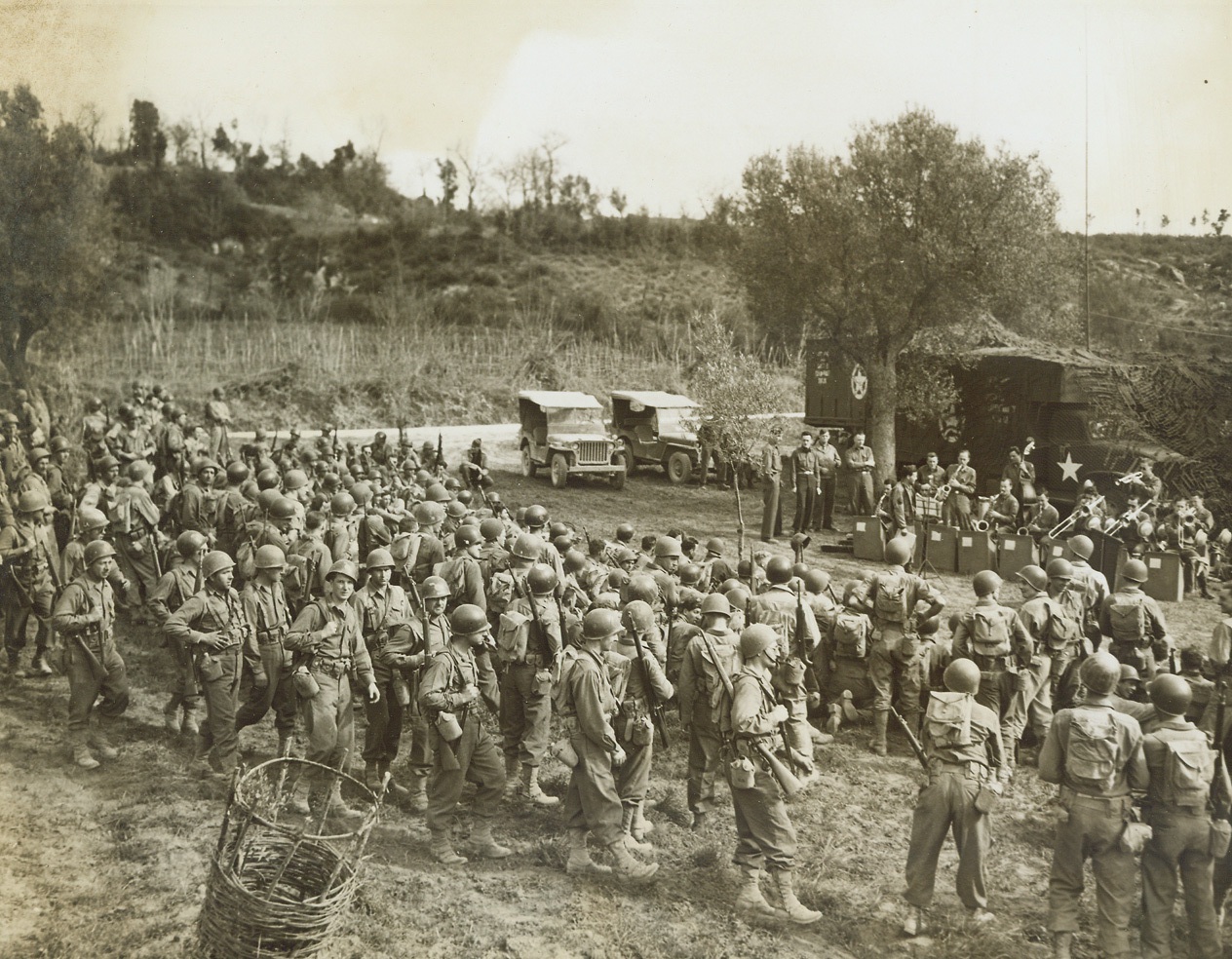 MOBILE RADIO STATION “GETS AROUND”, 4/19/1944. ITALY—War weary men and women of the Allied Fifth Army in Italy are mighty proud of their mobile radio station, officially known as the Fifth Army Mobile American Expeditionary Station, which gets it [sic] music and its “big time” programs to them wherever they are—in the front lines, or in rest camps. The hard working crew that moves the station’s ten-unit “circus caravan” of jeeps and trailers, and 2-1/2 ton trucks to various points in the combat area making sure that every group is reached at least once a day, have the moving operation down to a fine point. They can take the station down, move it 50 miles (which is the range of the transmitter), and set it up again, all within less than two hours. Here, a Fifth Army band set up before the station “somewhere in Italy,” furnishes a swing concert for Combat Engineers of the 337th Battalion, who call a halt to listen. At the same time, the music is broadcast by the station.Credit: Acme photo by Sherman Montrose for the War Picture Pool;