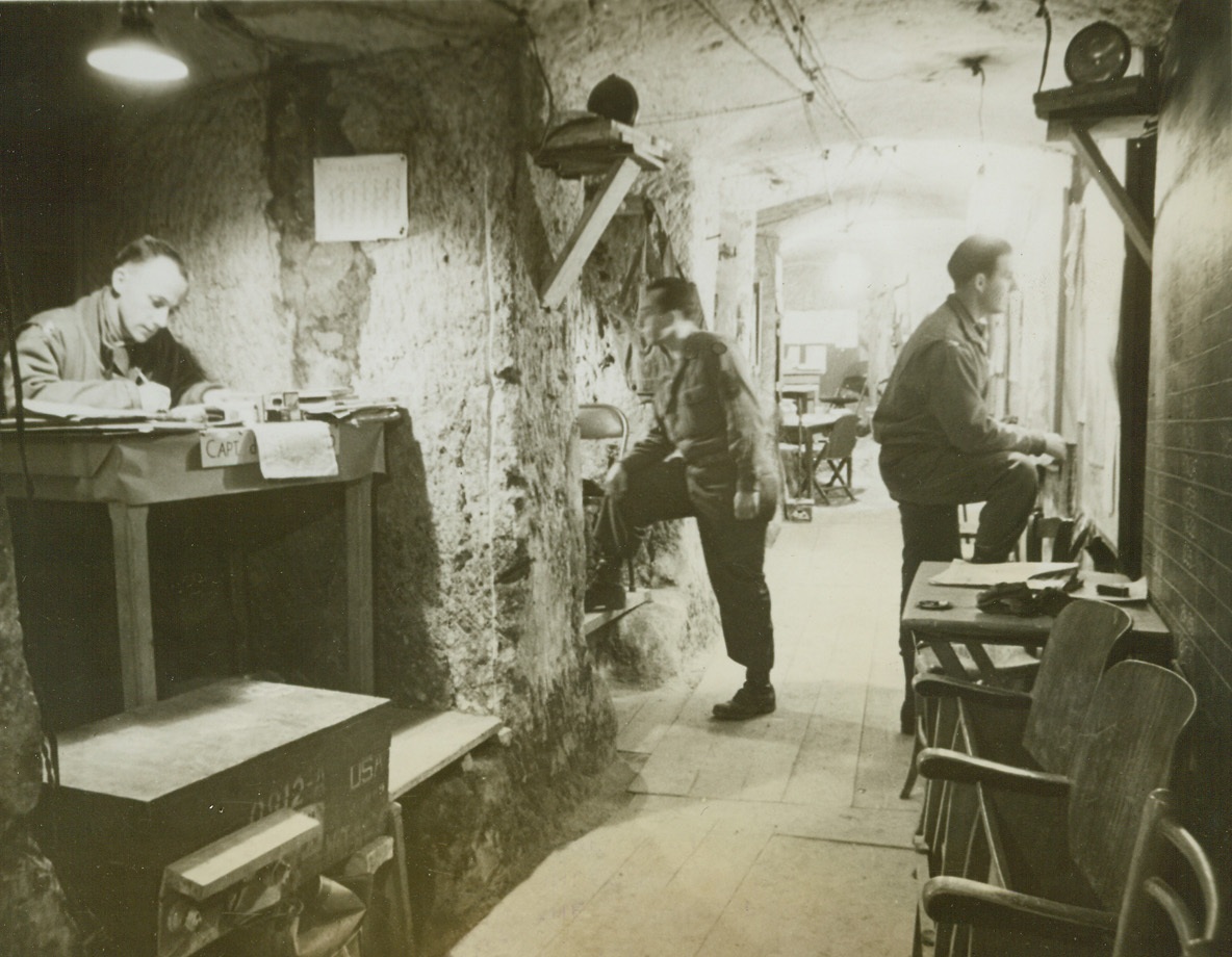 In the Tombs of the Ancient, 4/22/1944. NETTUNO, ITALY – When the Allied forces reached Nettuno, they found underground headquarters already built for them. The ancient catacombs make excellent living and working quarters for the men. The niches in the walls, formerly used to hold wine barrels, have been converted into bedrooms or offices. Note the calendar by the captain’s table, with the days carefully X’d out. Credit (Acme) (WP);